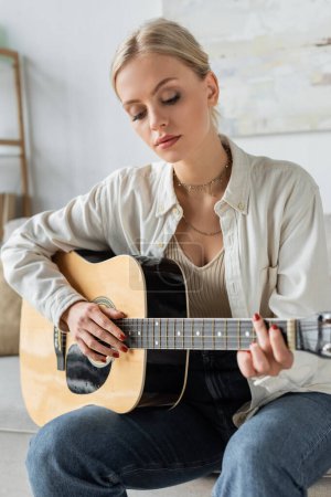 Foto de Young and blonde woman playing acoustic guitar while sitting on couch - Imagen libre de derechos