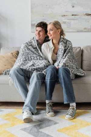 full length of happy young man and blonde woman covered in blanket sitting on couch 