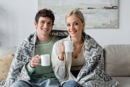 Photo for Happy young man and smiling blonde woman covered in blanket holding cups of tea while sitting on couch - Royalty Free Image
