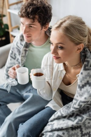 Photo for High angle view of young man and smiling woman covered in blanket holding cups of tea while sitting on couch - Royalty Free Image
