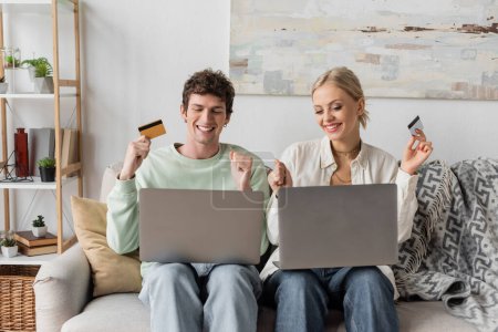 Foto de Happy young couple holding credit cards near laptops while doing online shopping - Imagen libre de derechos