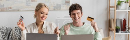 happy young couple holding credit cards near laptops while doing online shopping, banner 