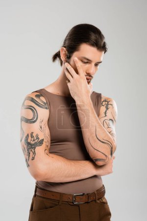 Photo for Long haired and tattooed man touching face while posing isolated on grey - Royalty Free Image