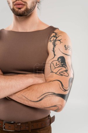 Cropped view of young bearded man with tattoo crossing arms isolated on grey 
