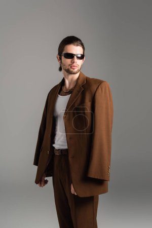 Fashionable man in sunglasses posing in brown suit isolated on grey 