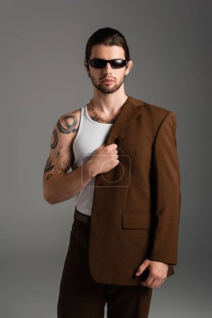 Bearded tattooed man in sunglasses holding jacket isolated on grey 