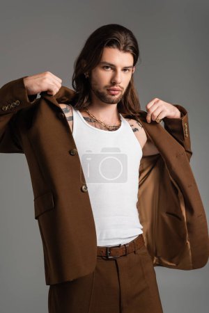 Trendy long haired man with tattoo wearing jacket isolated on grey 