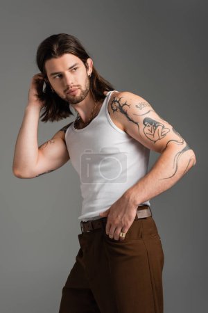 Photo for Trendy tattooed man in white sleeveless shirt adjusting hair isolated on grey - Royalty Free Image