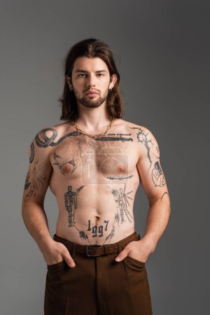 Long haired and tattooed man in pants posing isolated on grey 