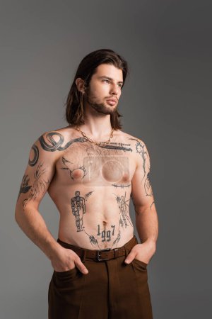 Muscular and tattooed man holding hands in pockets of pants isolated on grey 
