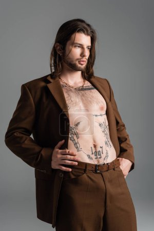 Shirtless and tattooed man in suit standing isolated on grey 