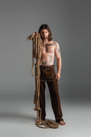 Foto de Barefoot and tattooed model holding rope on grey background - Imagen libre de derechos