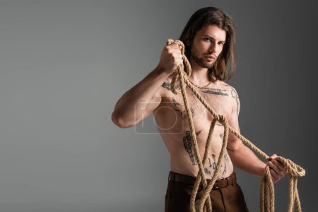 Young long haired model with tattoo on body holding rope isolated on grey 