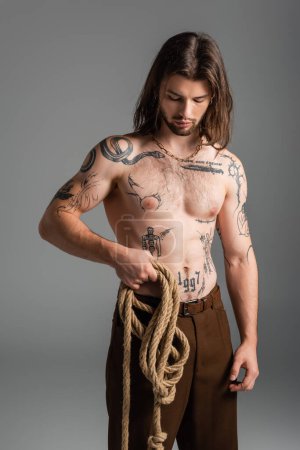 Foto de Long haired and tattooed model holding rope isolated on grey - Imagen libre de derechos
