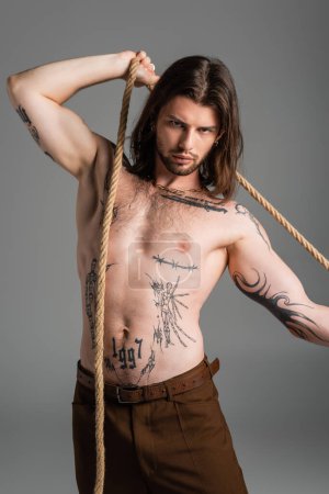 Foto de Shirtless tattooed model posing with rope and looking at camera isolated on grey - Imagen libre de derechos