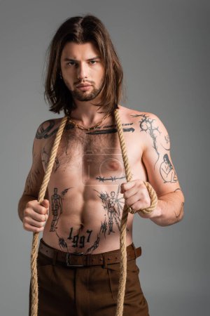 Photo for Sexy tattooed man holding rope and looking at camera isolated on grey - Royalty Free Image