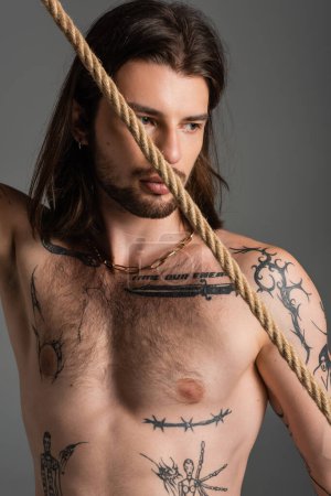 Shirtless tattooed man looking away near rope isolated on grey 