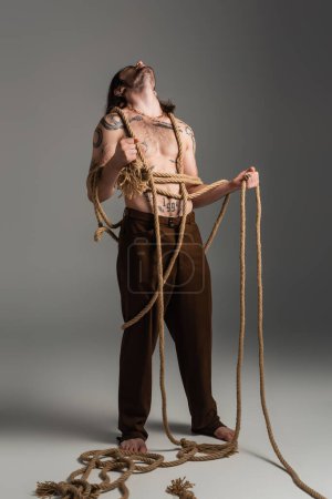 Log haired and tattooed model in pants holding rope on grey background 