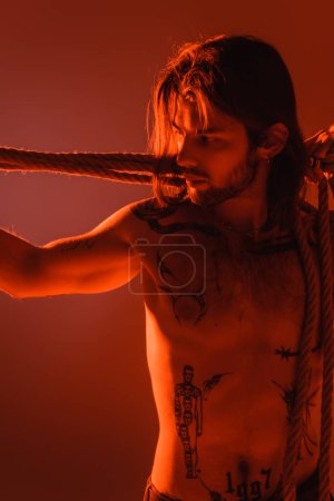 Long haired and tattooed man looking at rope isolated on red 