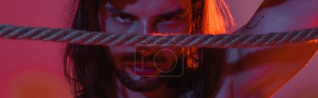 Photo for Long haired man looking at camera near rope on purple background with light, banner - Royalty Free Image