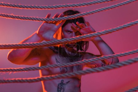 Sexy tattooed model posing near ropes on purple background red with light 