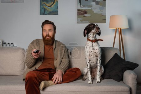 Bearded man in cardigan watching movie on couch near dalmatian dog at home 