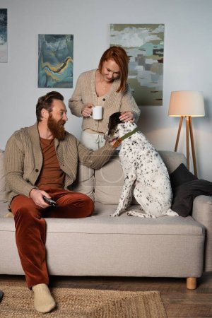 Cheerful man with remote controller and woman with cup petting dalmatian dog at home 