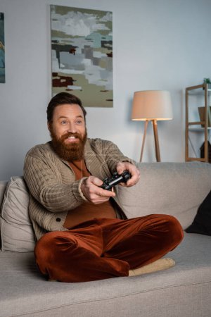 Téléchargez les photos : KYIV, UKRAINE - DECEMBER 16, 2022: Cheerful bearded man playing video game on couch at home - en image libre de droit