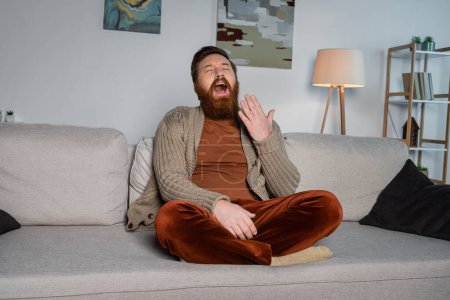 Photo for Exhausted bearded man yawning while sitting on couch at home - Royalty Free Image