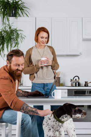 Fröhlich tätowierter Mann streichelt Dalmatiner-Hund neben Laptop und Ehefrau mit Tasse in Küche 