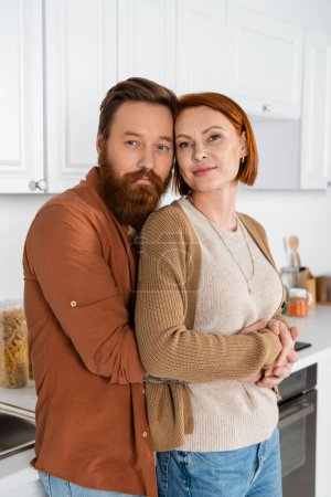 Bearded man hugging redhead wife in kitchen 