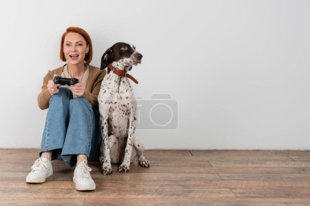 Photo for KYIV, UKRAINE - DECEMBER 16, 2022: Cheerful redhead woman playing video game near dalmatian dog at home - Royalty Free Image