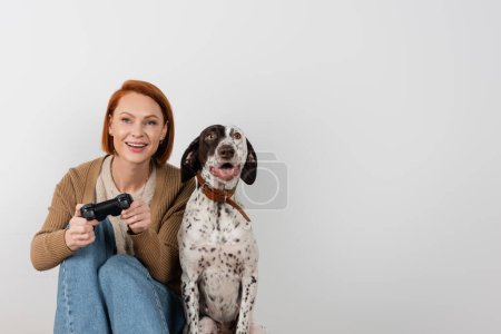Photo for KYIV, UKRAINE - DECEMBER 16, 2022: Smiling red haired woman playing video game near dalmatian dog - Royalty Free Image