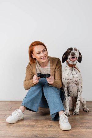 Photo for KYIV, UKRAINE - DECEMBER 16, 2022: Cheerful red haired woman holding joystick and looking at dalmatian dog at home - Royalty Free Image