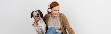 Smiling woman in wireless headphones hugging dalmatian dog at home, banner 