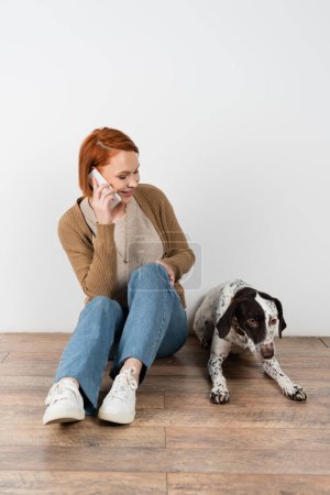 Smiling red haired woman talking on smartphone and looking at dalmatian dog on floor 