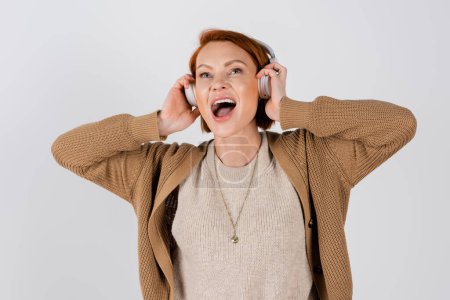 excited and red haired woman in headphones singing isolated on grey 