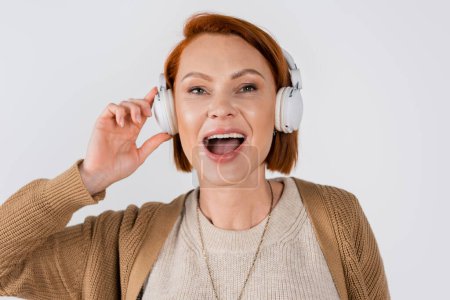 Photo for Portrait of red haired woman singing while using headphones isolated on grey - Royalty Free Image
