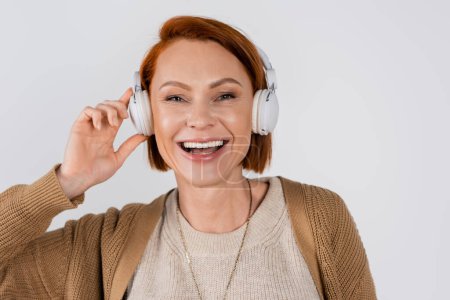 Photo for Portrait of cheerful red haired woman listening music in headphones isolated on grey - Royalty Free Image