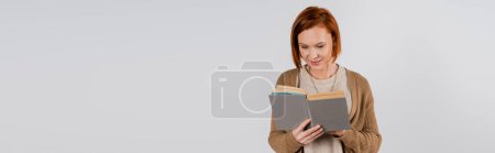 Adult redhead woman reading book isolated on grey with copy space, banner 