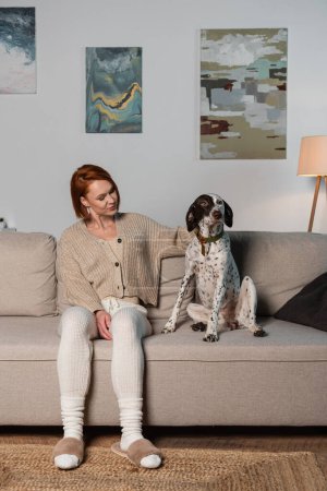 Mujer pelirroja en chaqueta mirando al perro dálmata en el sofá 