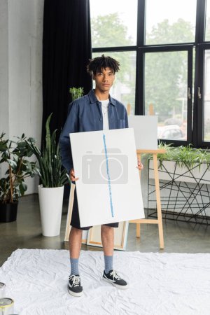 Young african american artist holding painting on cloth in studio 