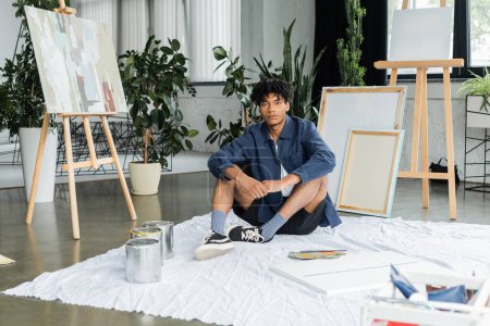 Artiste afro-américain regardant la caméra tout en étant assis près de peintures et de toile sur le sol dans l'atelier 
