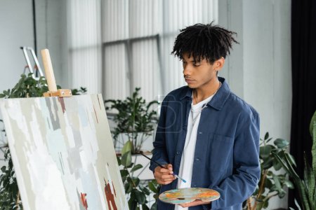 Young african american artist holding paintbrush and palette near painting in studio 
