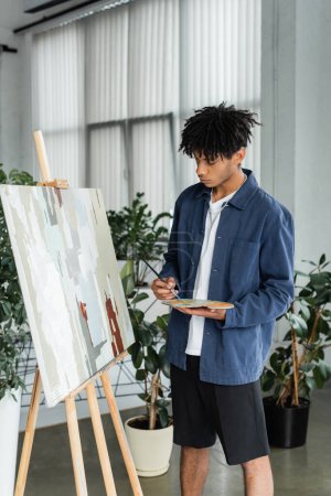 Artista afroamericano sosteniendo paleta y pincel cerca de dibujar en caballete 