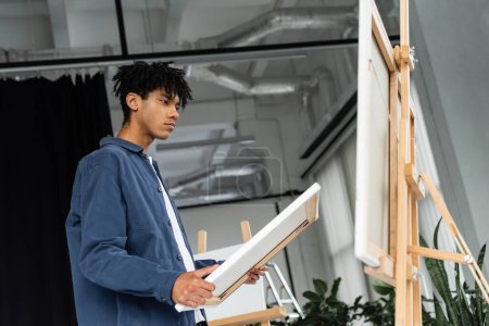 Low angle view of african american artist holding canvas near easel in studio 