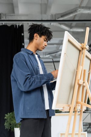 Foto de Side view of young african american artist holding paintbrush near canvas in studio - Imagen libre de derechos