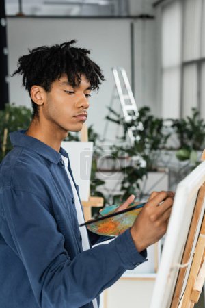 Artista afroamericano pintando sobre lienzo borroso y sosteniendo paleta en estudio 