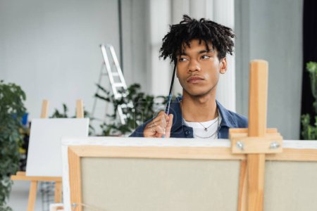 Photo for Pensive african american artist holding paintbrush near blurred canvas on easel in studio - Royalty Free Image