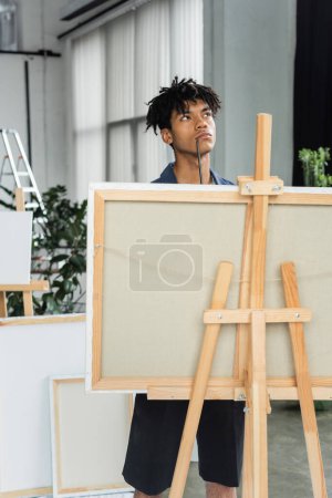 Pensativo artista afroamericano con pincel de pie cerca de la lona en el estudio